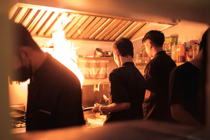 Brigade de cuisine au travail devant une flamme vive, sous une hotte d'extraction professionnelle.