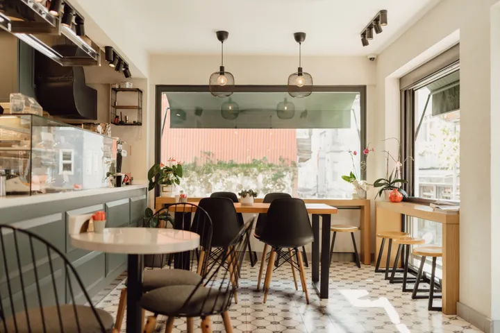 Salle intérieure d'un coffee shop avec tables, chaises et grande baie vitrée baignée de lumière naturelle.