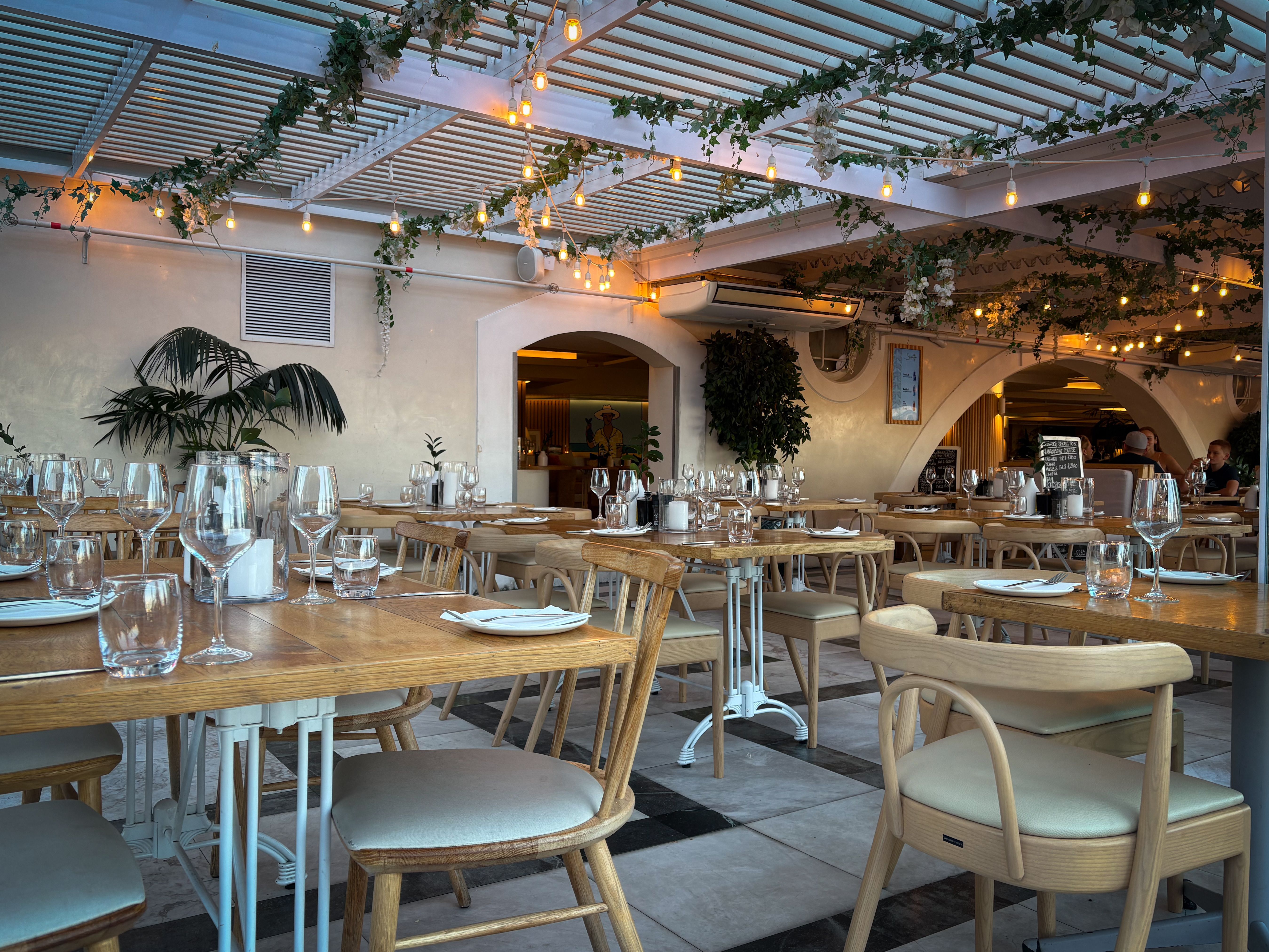 Terrasse de restaurant aménagée avec tables en bois, guirlandes lumineuses et végétation, en fin de journée.